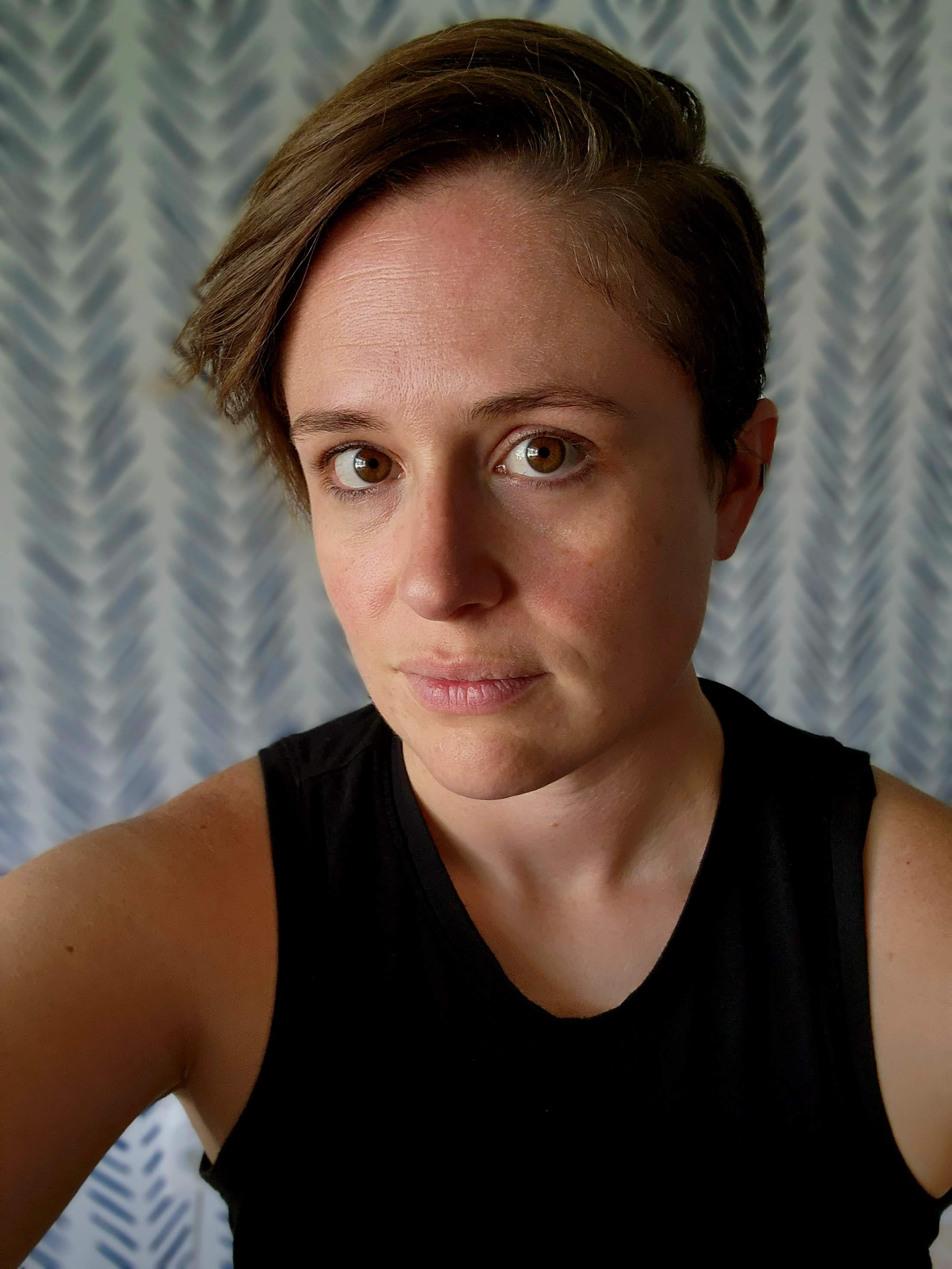 Image with a woman with short, brown hair in an undercut smiles at the camera with a blurred background