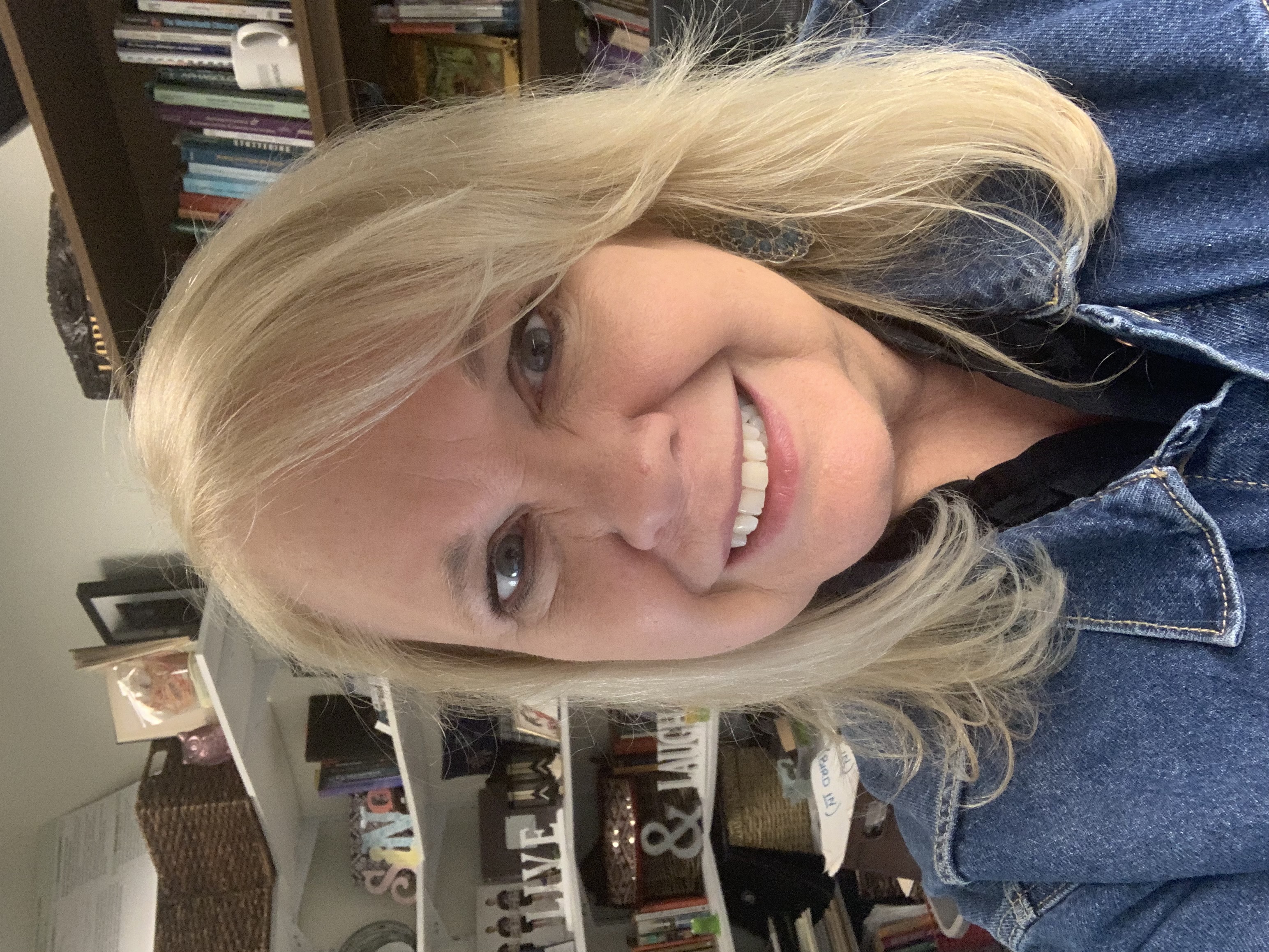 An image with a woman with blonde hair in a blue, denim shirt smiles at the camera with bookshelves in the background