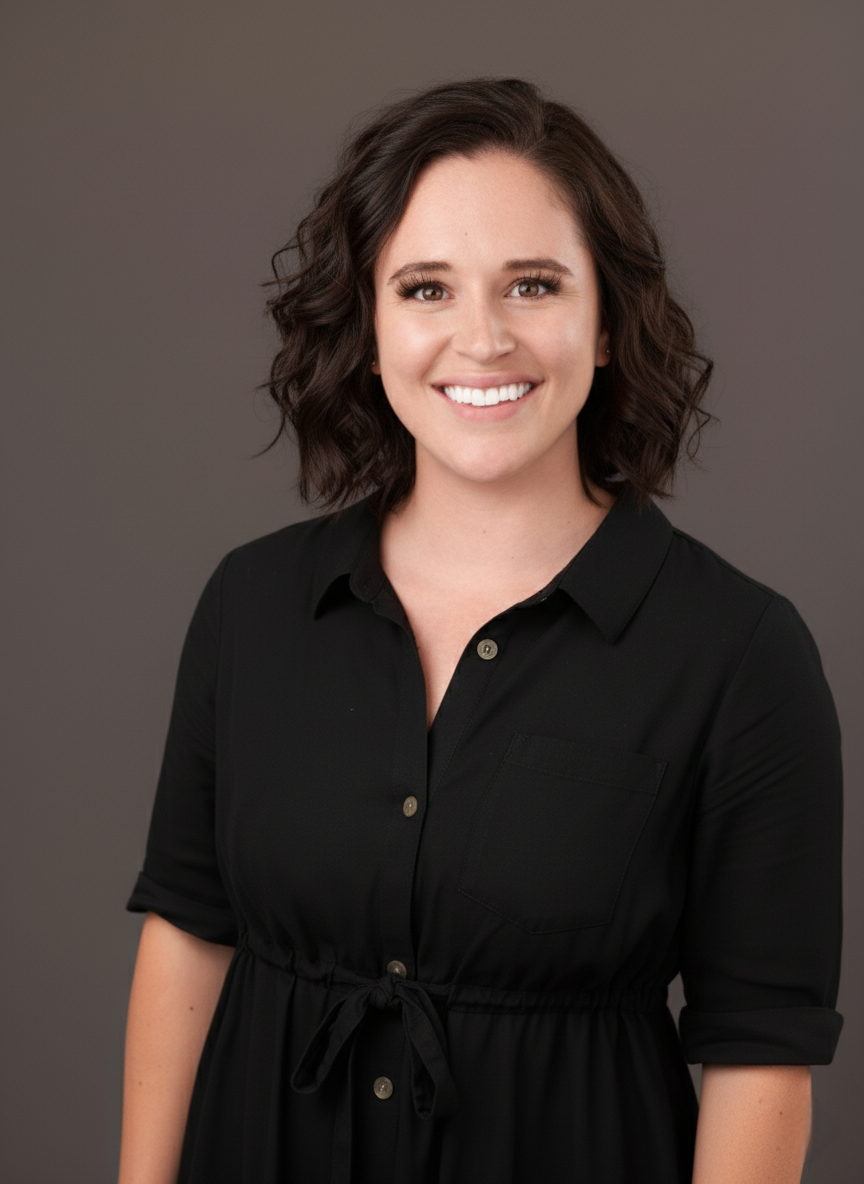 An image with a caucasian woman with shoulder-length brown, curly hair smiling at the camera. She is wearing black and is standing against a grey background