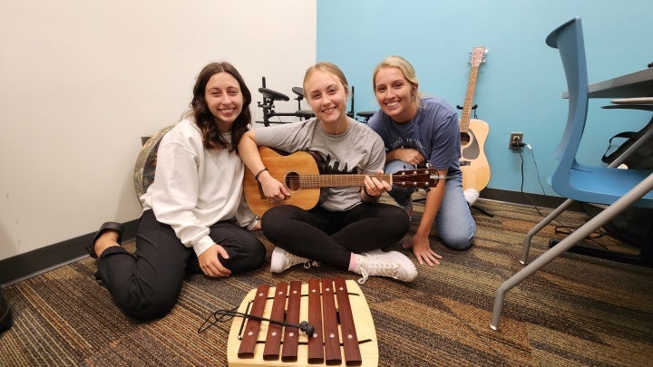 Three students sit together in a music therapy clinic space. One holds a guitar, with two others sitting beside them, all smiling. An instrument rests on the floor in front of them, adding to the relaxed, welcoming atmosphere.