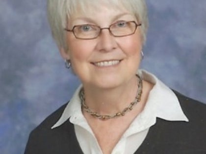 An image of a caucasian woman with short white hair and glasses against a blue background